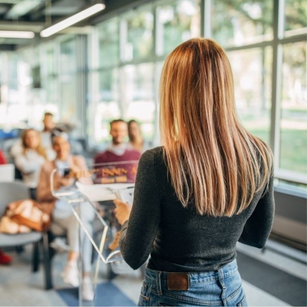 Jeune femme blonde de dos face à un public lors d'une présentation oral